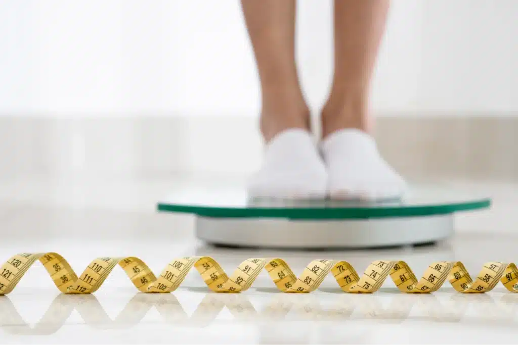 A person in white socks stands on a scale. A yellow tape measure is placed in the foreground.